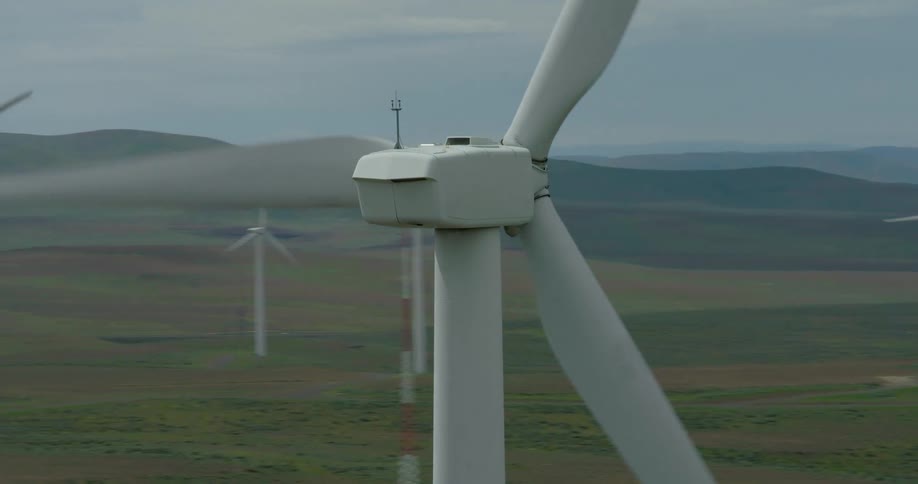 An aerial close-up orbits a spinning wind turbine, revealing a large wind farm spread across rolling green hills