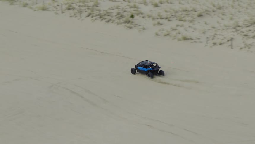 Slow motion aerial footage follows a blue dune buggy as it drives across expansive sand dunes