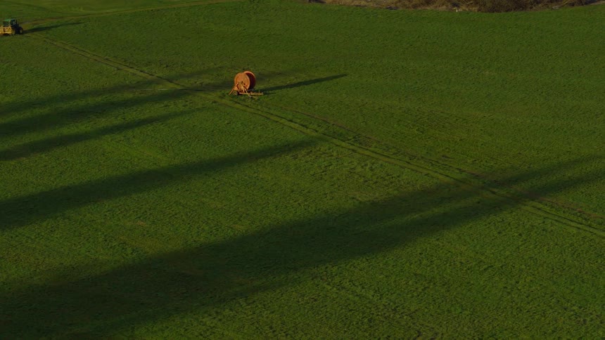 Long shadows stretch across a lush green field where a single hay bale sits, seen from an aerial perspective at sunrise