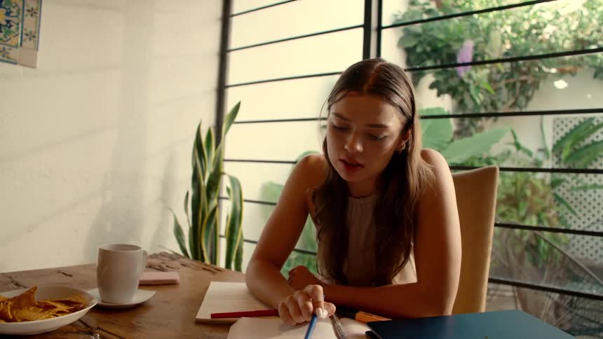 A pensive young woman studies at home, writing in her notebook and drinking from a mug in a cozy, sun-drenched room