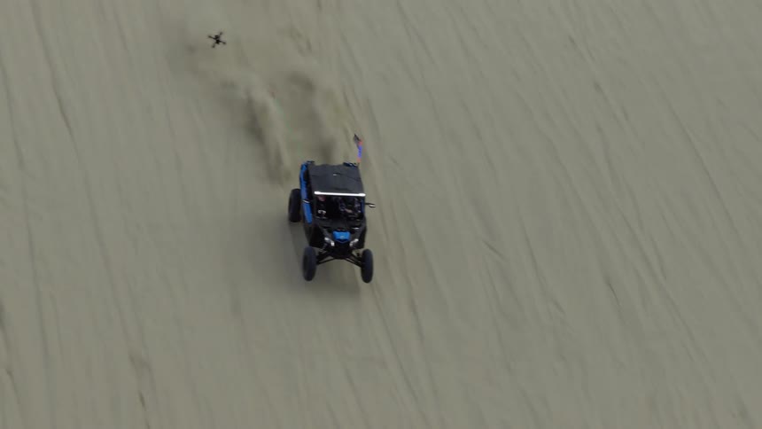 An aerial shot follows a blue dune buggy as it kicks up sand while driving across a vast desert dune