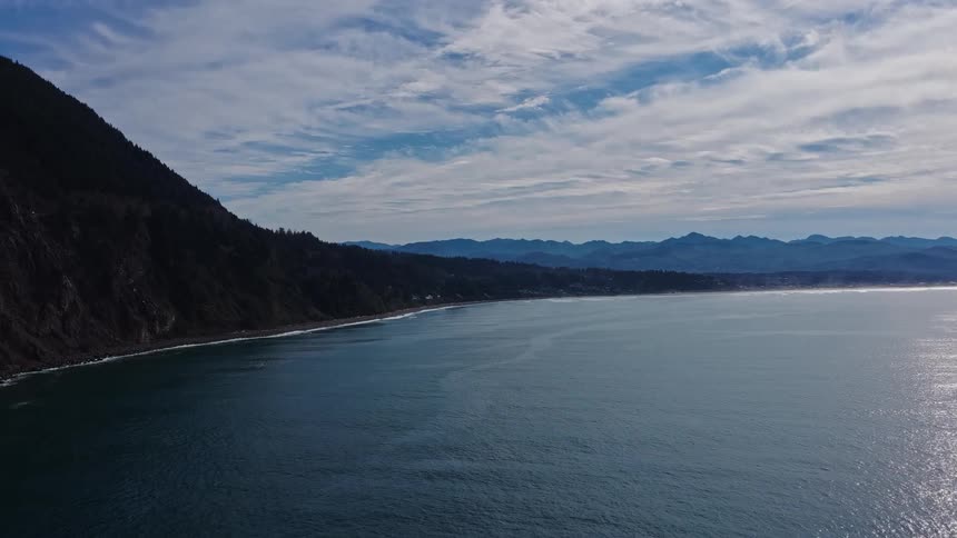 A stunning aerial view of the sun-kissed ocean slowly pans to reveal a rugged, mountainous coastline under a beautiful sky