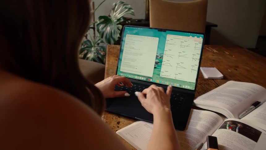 Over the shoulder shot of a female student studying mathematical graphs and taking notes on her laptop at home
