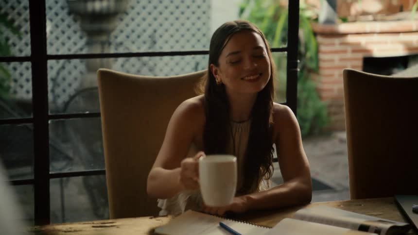 A smiling young woman sips her morning coffee in slow motion while studying at a wooden table and happily engages in conversation