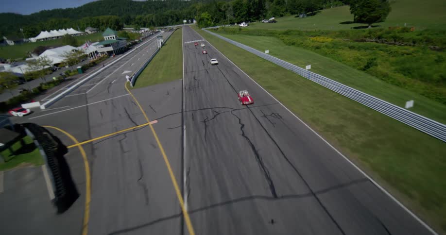 A dynamic aerial shot flies over a race track, following a pack of colorful vintage cars during a race