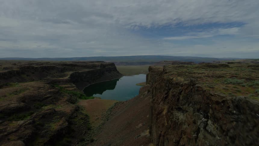 A majestic aerial view retreats slowly over the rugged cliffs of a vast canyon, revealing a serene lake below under a cloudy sky
