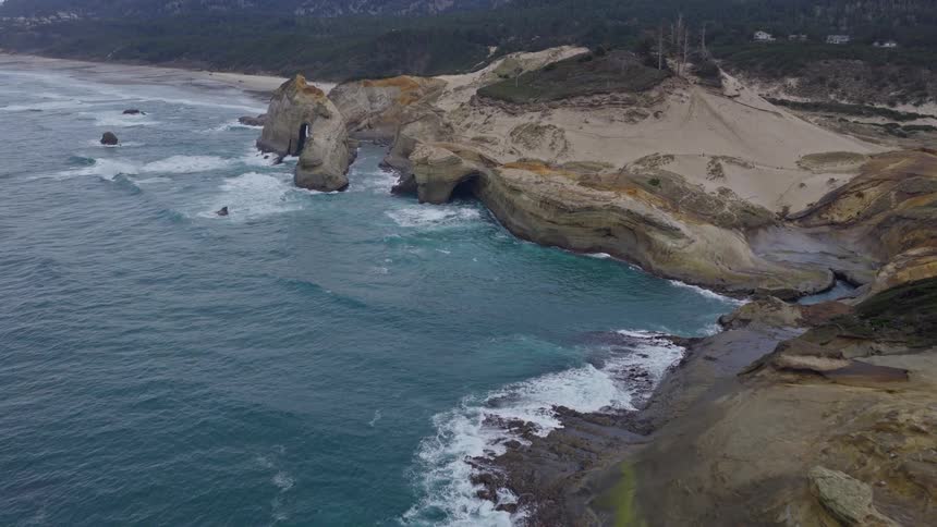 An aerial view reveals a rugged and dramatic coastline with sea stacks, natural arches, and sand dunes as waves crash ashore