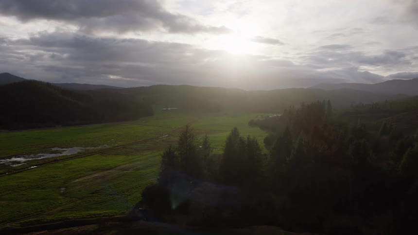 A slow aerial pullback reveals a beautiful sunlit valley surrounded by mountains and a dense evergreen forest