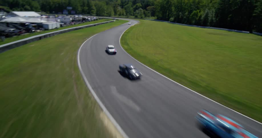 A thrilling, high-speed FPV drone follows several vintage race cars as they navigate a turn on a professional racetrack