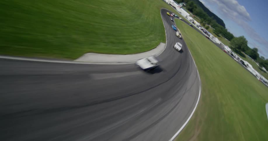 High-speed aerial footage tracks a pack of colorful vintage sports cars as they speed around a winding racetrack on a sunny day