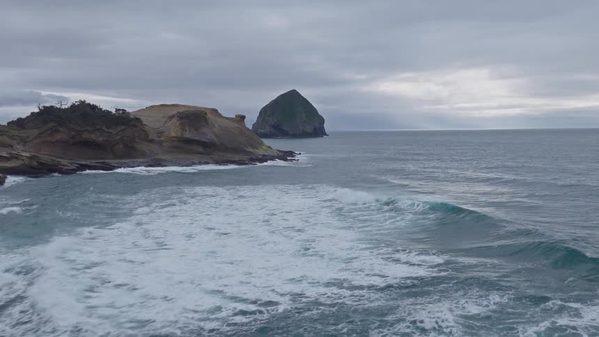 A dramatic slow-motion aerial shot flies over choppy ocean waves towards the rugged cliffs and sea stacks of the Oregon coast on a cloudy day