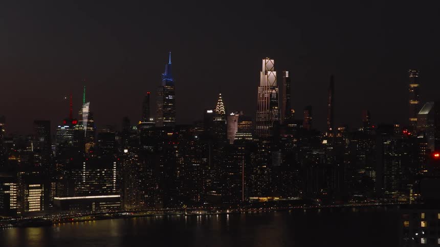 Aerial pull-back shot of the iconic Manhattan skyline with brightly lit skyscrapers reflecting in the East River at night
