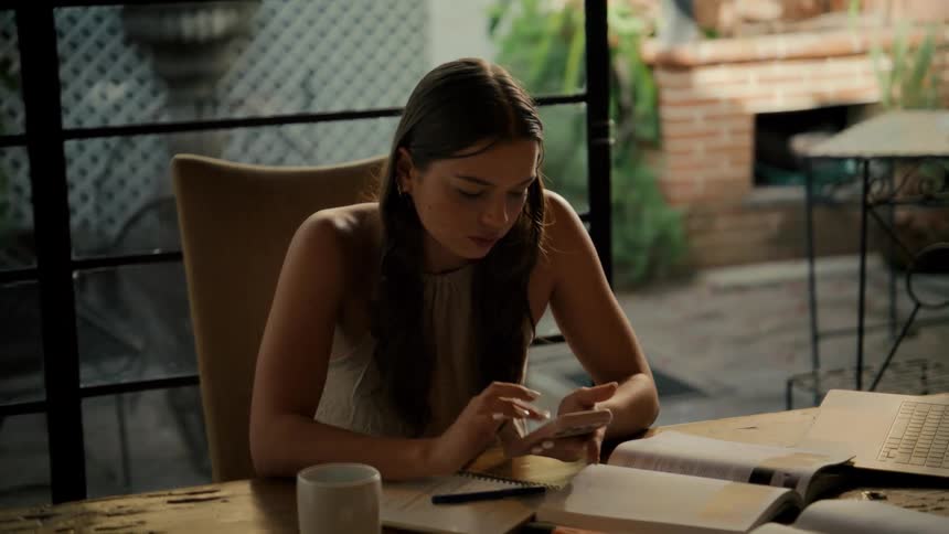 A young woman sits at her desk with books and a laptop, using her smartphone to assist with her studies in a softly lit room