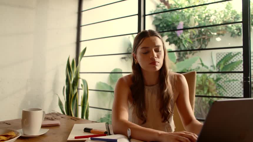 A stressed young student anxiously works on her laptop at a sunlit table, her worried expression slowly turning into relief