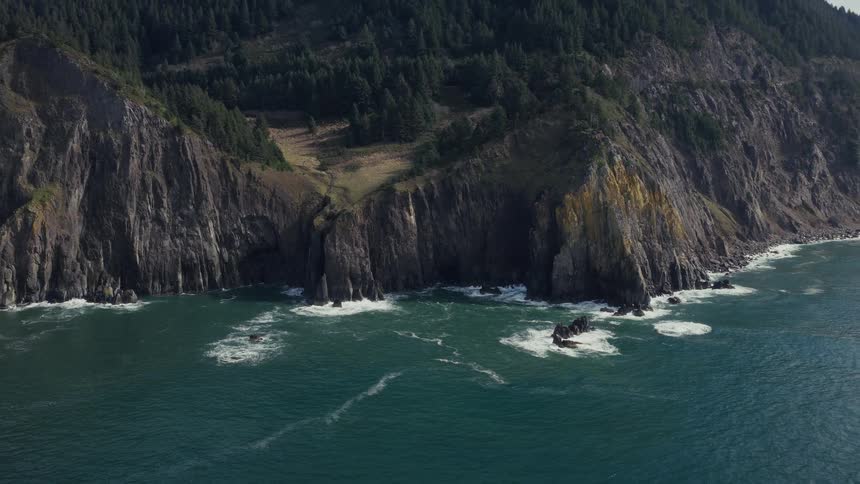 A slow aerial pullback reveals a rugged coastline where a vast, forest-covered mountain meets the powerful ocean waves
