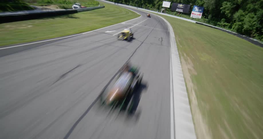 A dynamic aerial shot follows vintage open-wheel race cars speeding around a professional racetrack on a sunny day