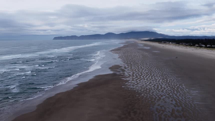Drone gracefully descends over a vast, moody beach, revealing intricate patterns in the wet sand at low tide on an overcast day
