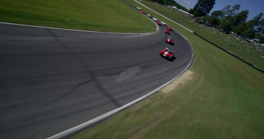 An exhilarating FPV drone shot follows a pack of classic sports cars racing around a scenic track on a sunny day