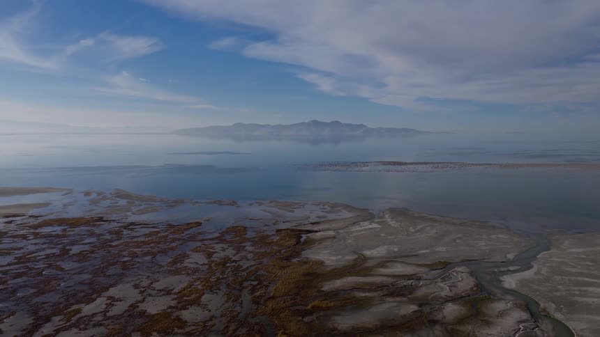 A vast aerial view of a marshy shoreline meeting a calm lake with a distant mountain island perfectly reflected on the water