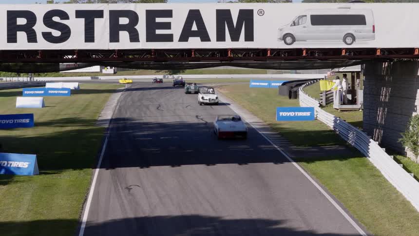 Classic race cars speed down a sunny racetrack under a branded Airstream bridge