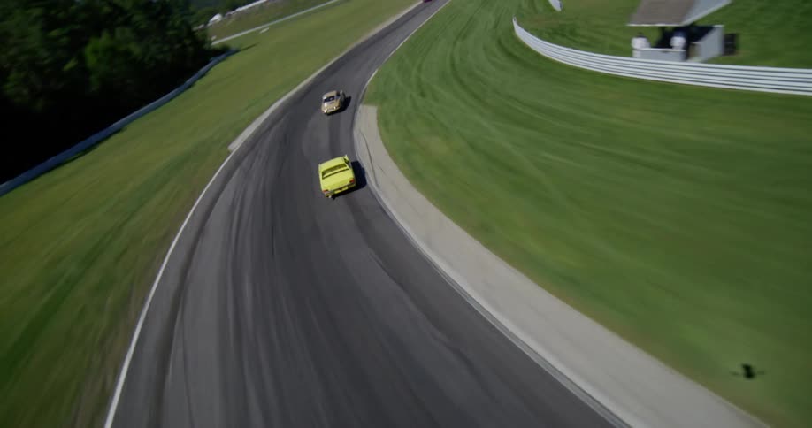 Dynamic aerial view follows vintage race cars speeding around a scenic, green racetrack on a sunny day