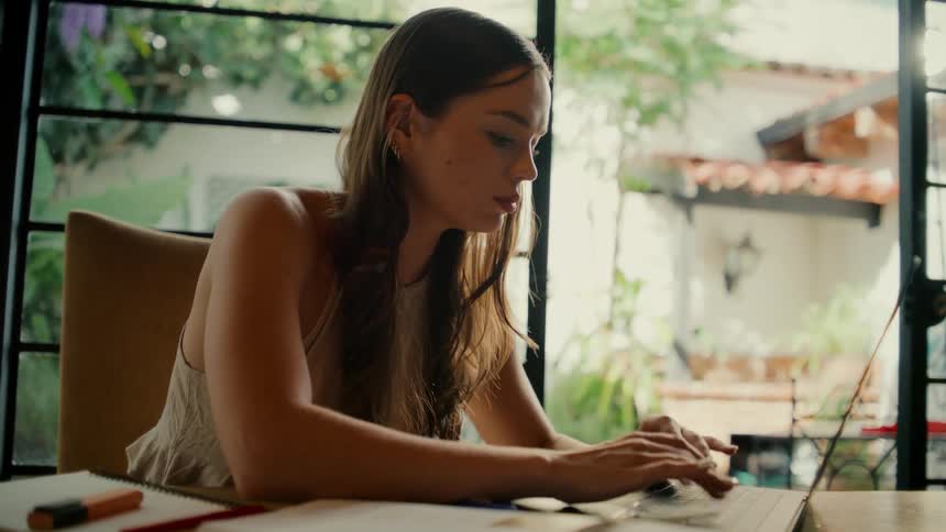 A young woman studies at home, highlighting notes before opening her laptop to work by a sunny window