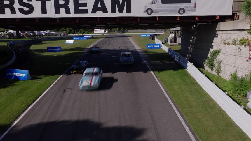 A static aerial shot shows various race cars speeding down a sunlit racetrack and passing under a bridge during a race