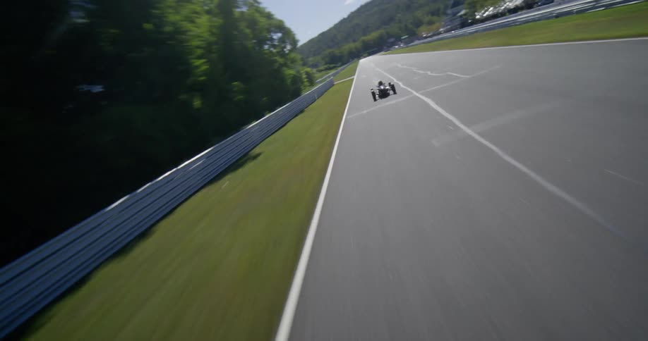 A dynamic aerial view follows a single-seater race car as it accelerates down a long straight on a sunny racetrack