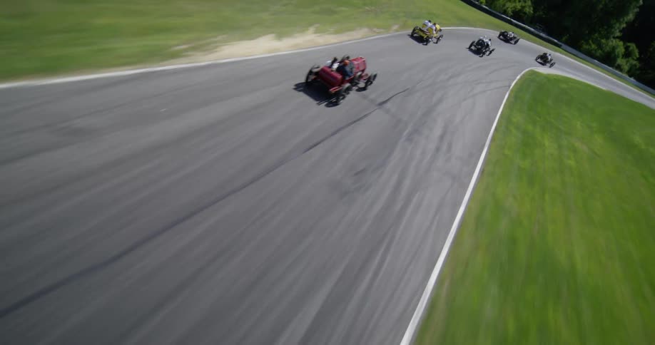 Dynamic aerial shot follows classic race cars speeding around a winding track in the scenic countryside