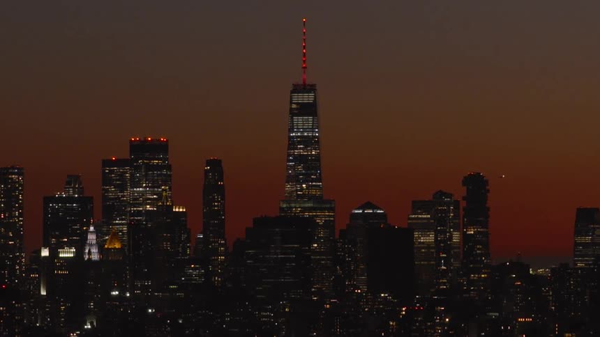 The iconic Lower Manhattan skyline glows at dusk as the One World Trade Center's spire changes from red to purple and blue