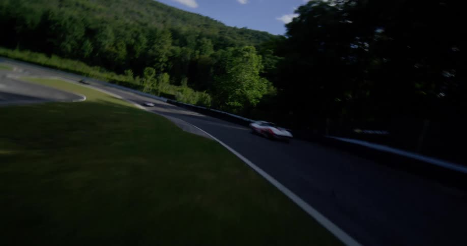 A thrilling low-level aerial shot follows vintage race cars as they speed around a winding track on a sunny day