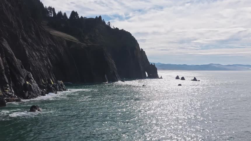An epic aerial shot glides between a towering sea stack and a sheer cliff face to reveal the spectacular and rugged Oregon coastline