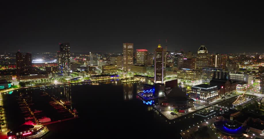 A slow aerial retreat reveals the sparkling Baltimore city skyline and Inner Harbor with beautiful lights reflecting in the water at night