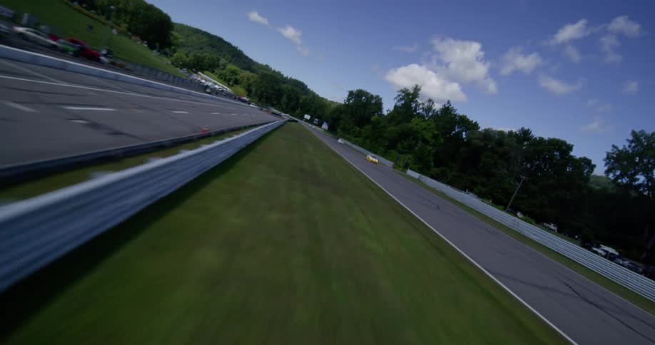 An aerial view flies over the grandstands and down the main straightaway of a racetrack as race cars speed by