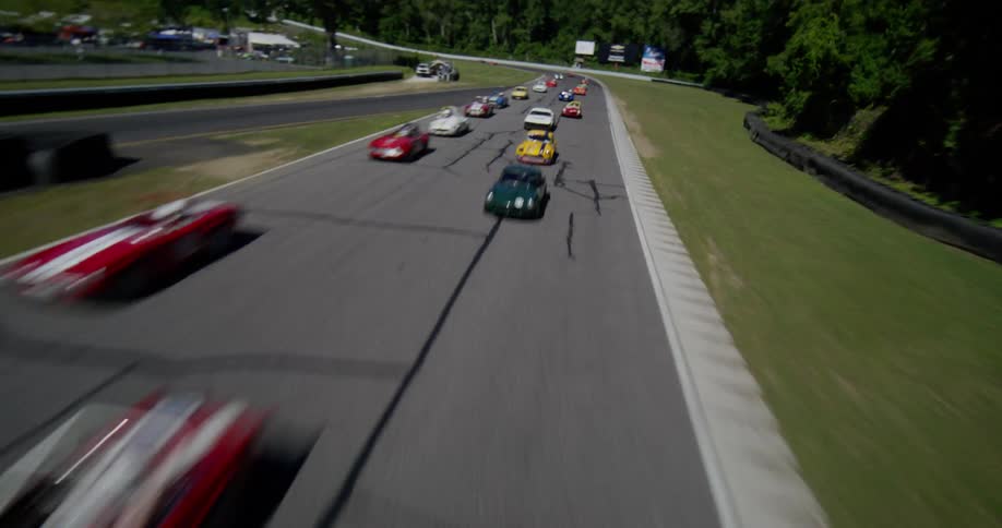 A high-speed drone flies over the starting line of a racetrack, then follows a pack of colorful vintage sports cars as they compete