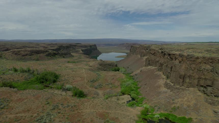 A slow aerial push reveals a hidden lake and small oasis within a vast, arid canyon under a partly cloudy sky