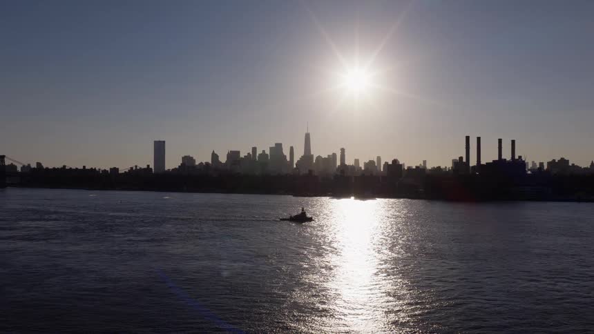 A tugboat cruises along the river with the iconic New York City skyline silhouetted by the bright sun