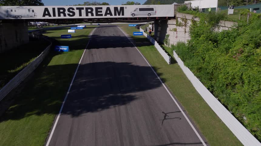 High angle aerial of vintage race cars speeding down a straightaway before the drone flies under a sponsored bridge