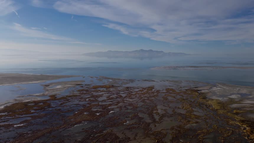 A sweeping aerial view slowly pulls back over the icy wetlands on the shore of the Great Salt Lake, revealing the vast, calm waters and a distant mountain island