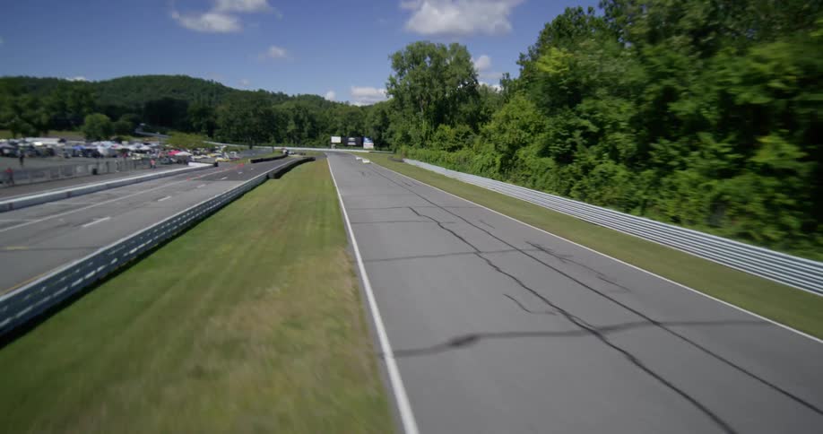 A fast-moving aerial shot flies over a creek and onto a racetrack, chasing two white sports cars as they race down the straightaway on a sunny day