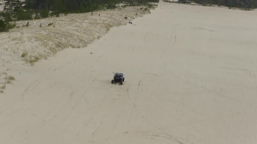 An aerial shot follows a blue UTV as it drifts in circles on a sand dune before driving across the vast sandy landscape