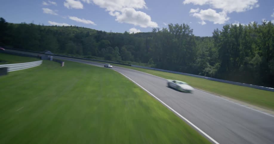 Dynamic aerial footage chases multiple colorful sports cars speeding around a winding race track in the countryside on a sunny day