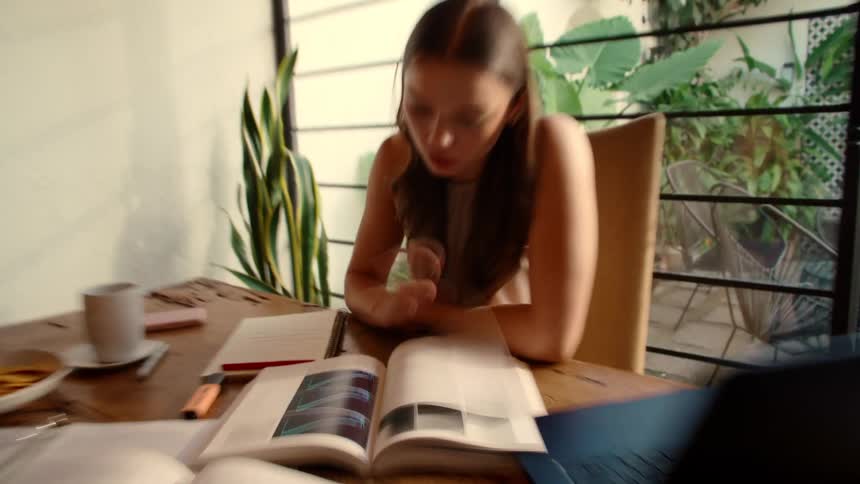 A young woman studies intensely at a wooden desk, surrounded by books, a laptop, and snacks, filmed with a dizzying camera effect