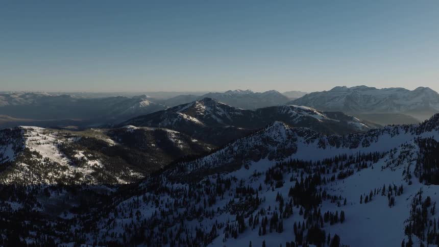 A serene aerial perspective of sun-kissed snowy peaks and forested valleys in a vast mountain range under a clear blue sky
