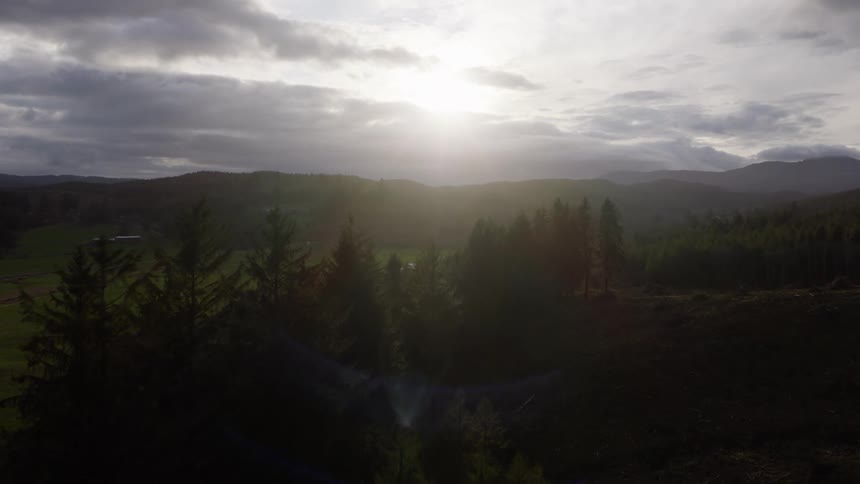 A slow aerial view flies over a backlit forest towards the bright sun breaking through dramatic clouds over a rural valley