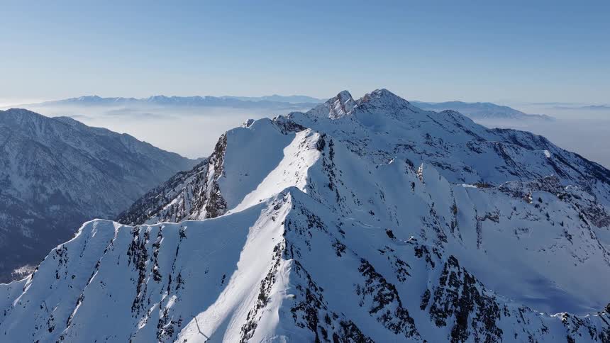 An epic aerial view pulls back along a sharp, snow-covered mountain ridge on a bright, sunny day