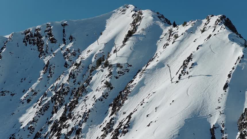 An aerial drone slowly pulls back to reveal the impressive scale of a jagged, snow-covered mountain peak against a clear blue sky