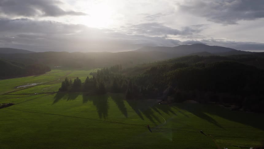 A dramatic aerial shot pulls back to reveal a vast rural landscape of green fields being watered by sprinklers as the sun breaks through clouds, casting long shadows from the forested hills