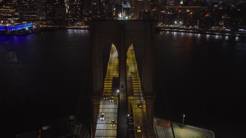 A high-angle static shot of traffic flowing over the illuminated Brooklyn Bridge at night with the New York City skyline in the background