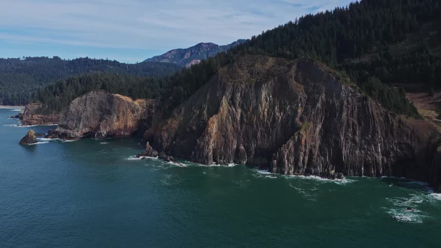A slow aerial pullback reveals the stunning and rugged coastline where majestic, tree-covered cliffs meet the deep blue ocean